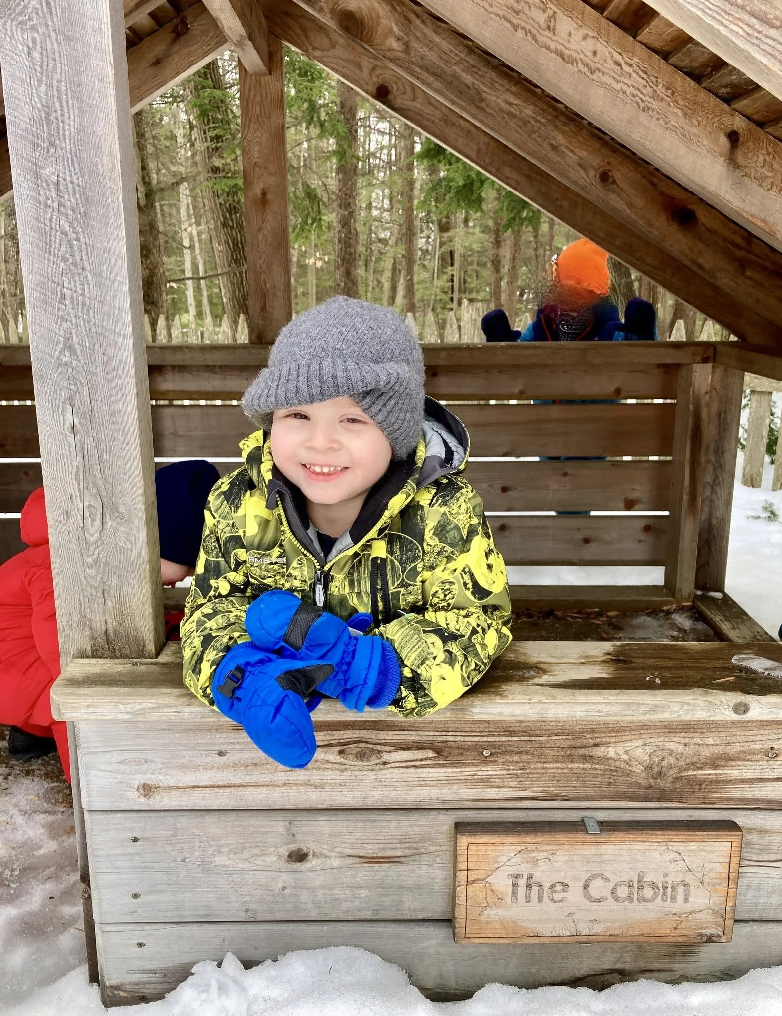 A young boy smiling and wearing a gray knit hat and yellow jacket with black patterns, leaning on a wooden structure labeled 'The Cabin' in snowy outdoor setting. Behind him, a person in an orange hood and black gloves is visible, surrounded by trees.