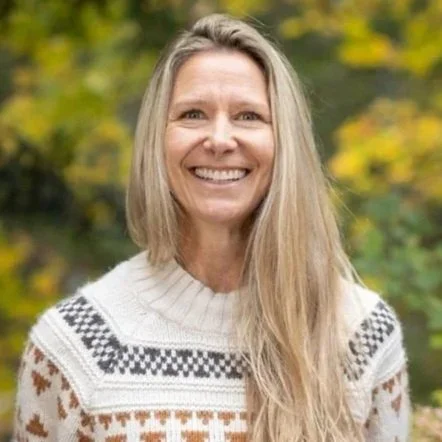 A smiling woman with long blonde hair standing outdoors with blurred green and yellow trees in the background.