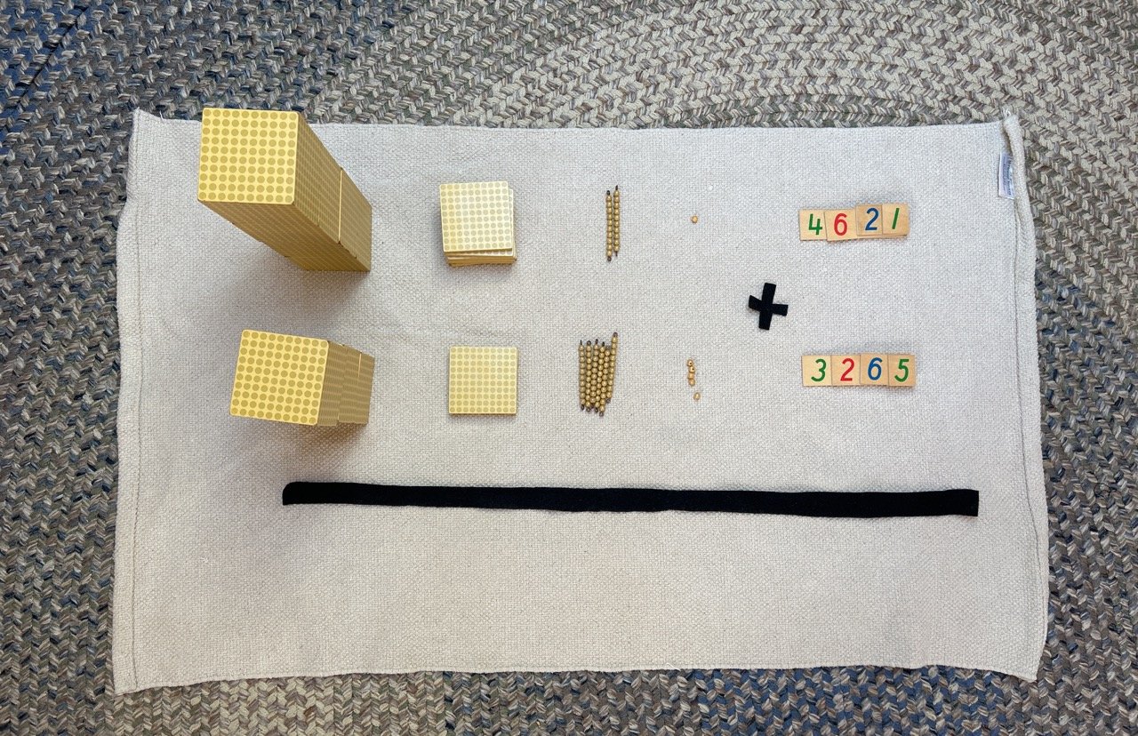 Educational math materials on a white cloth, including stacks of gold Lego blocks, small blocks, and various colored number tiles, with a black cloth strip at the bottom, on a textured gray background.