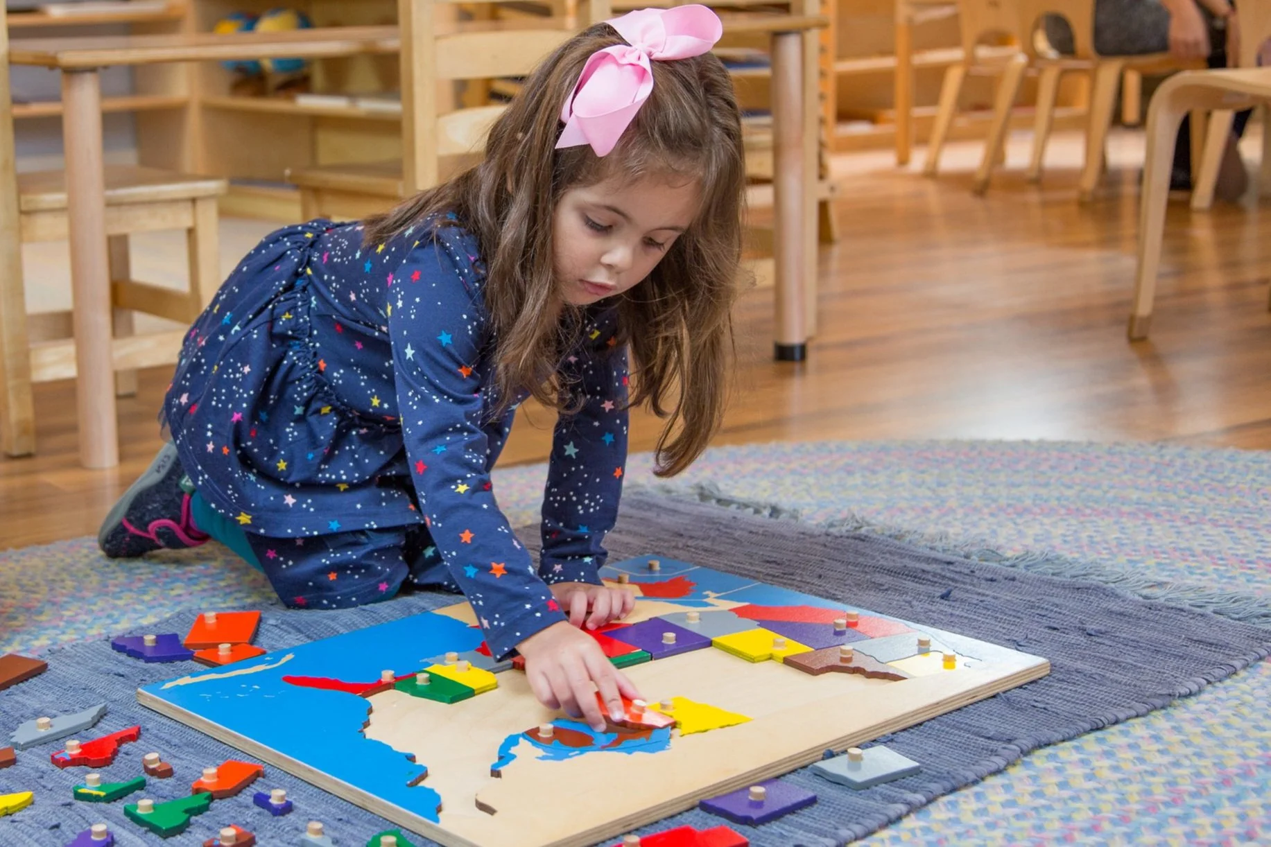 A young girl with long brown hair, wearing a navy dress with colorful star patterns and a pink bow headband, kneels on a carpet playing with a wooden puzzle map of the United States, surrounded by puzzle pieces, in a classroom with wooden furniture.