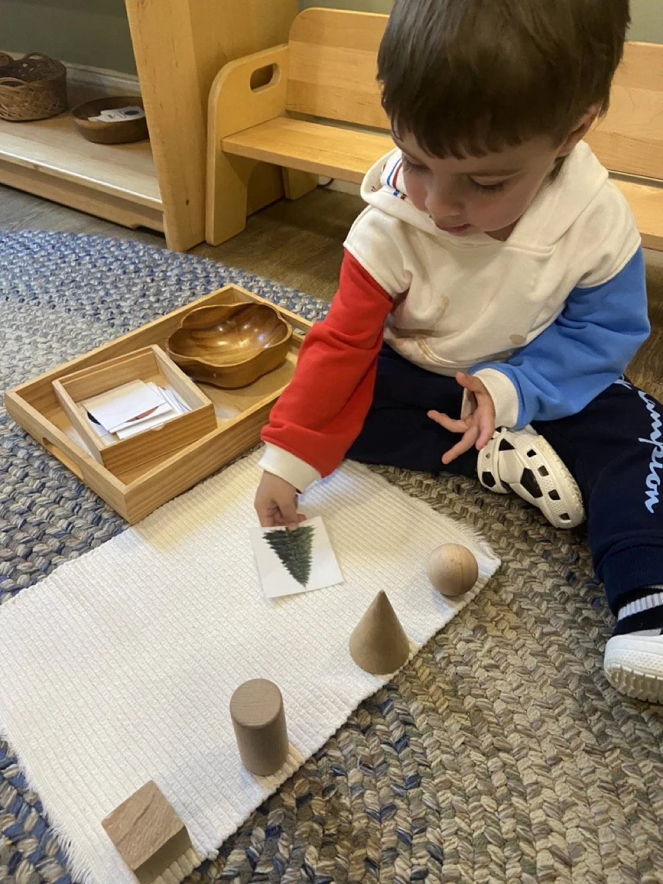 A young boy playing with wooden blocks on a white cloth, arranging shapes like a cone, sphere, and cube, with a picture of a Christmas tree nearby.