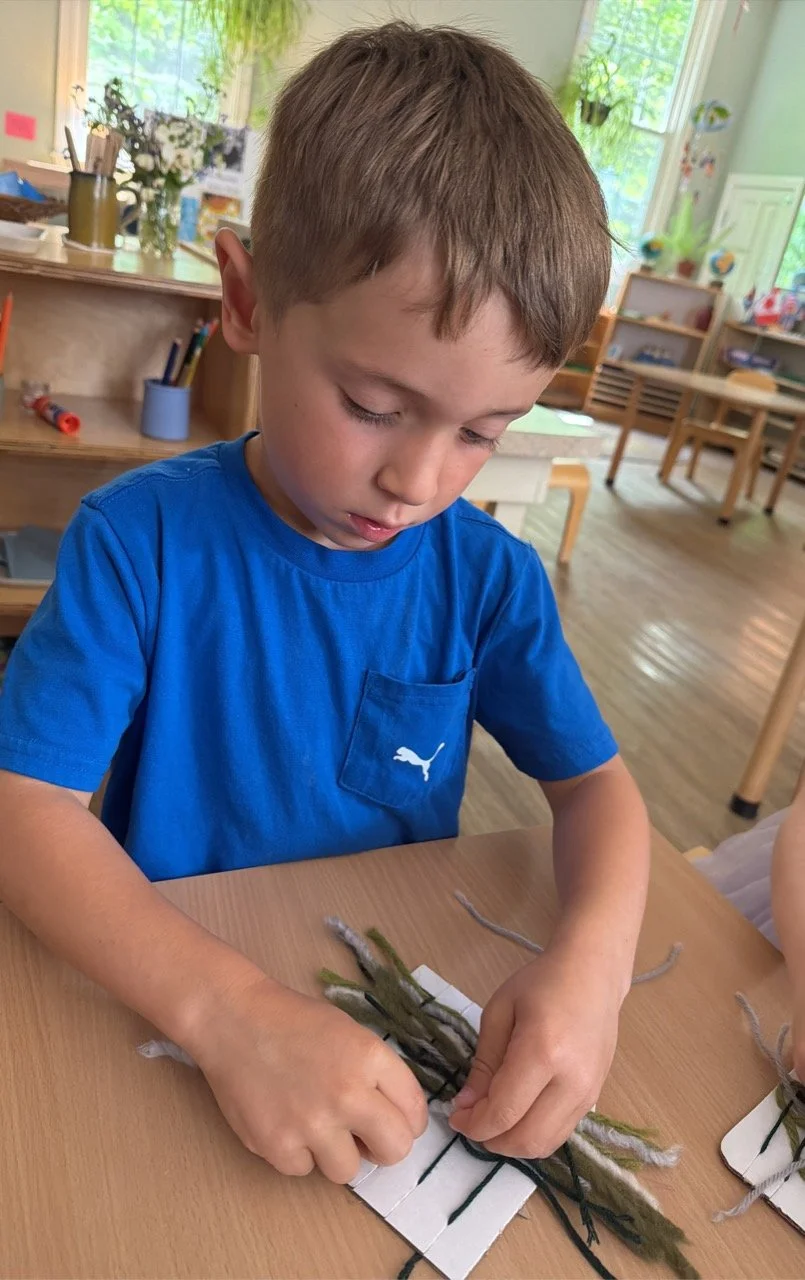 A young boy in a blue t-shirt working on a craft project with yarn on a wooden table in a classroom filled with natural light.