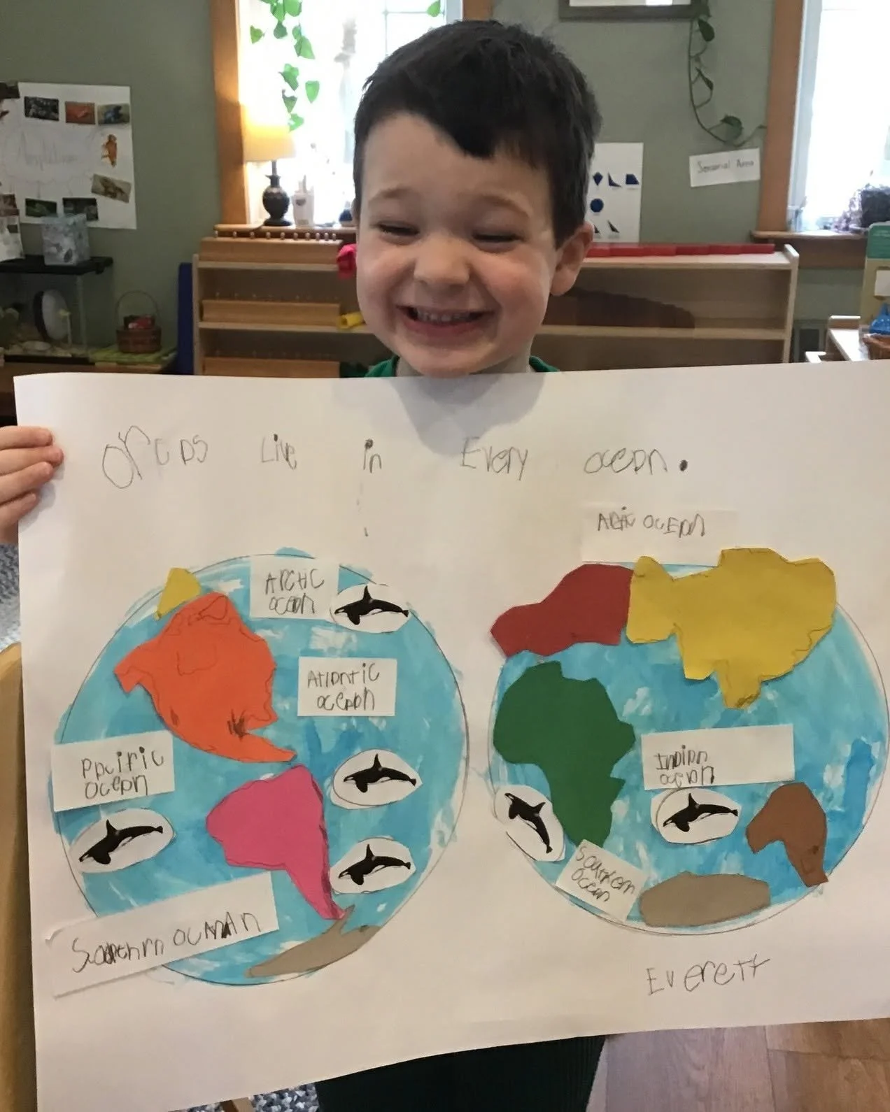 A young boy holding a poster displaying a colorful world map. The map is labeled with ocean names and features drawings of orcas. The boy is smiling and standing in a room with bookshelves and plants in the background.