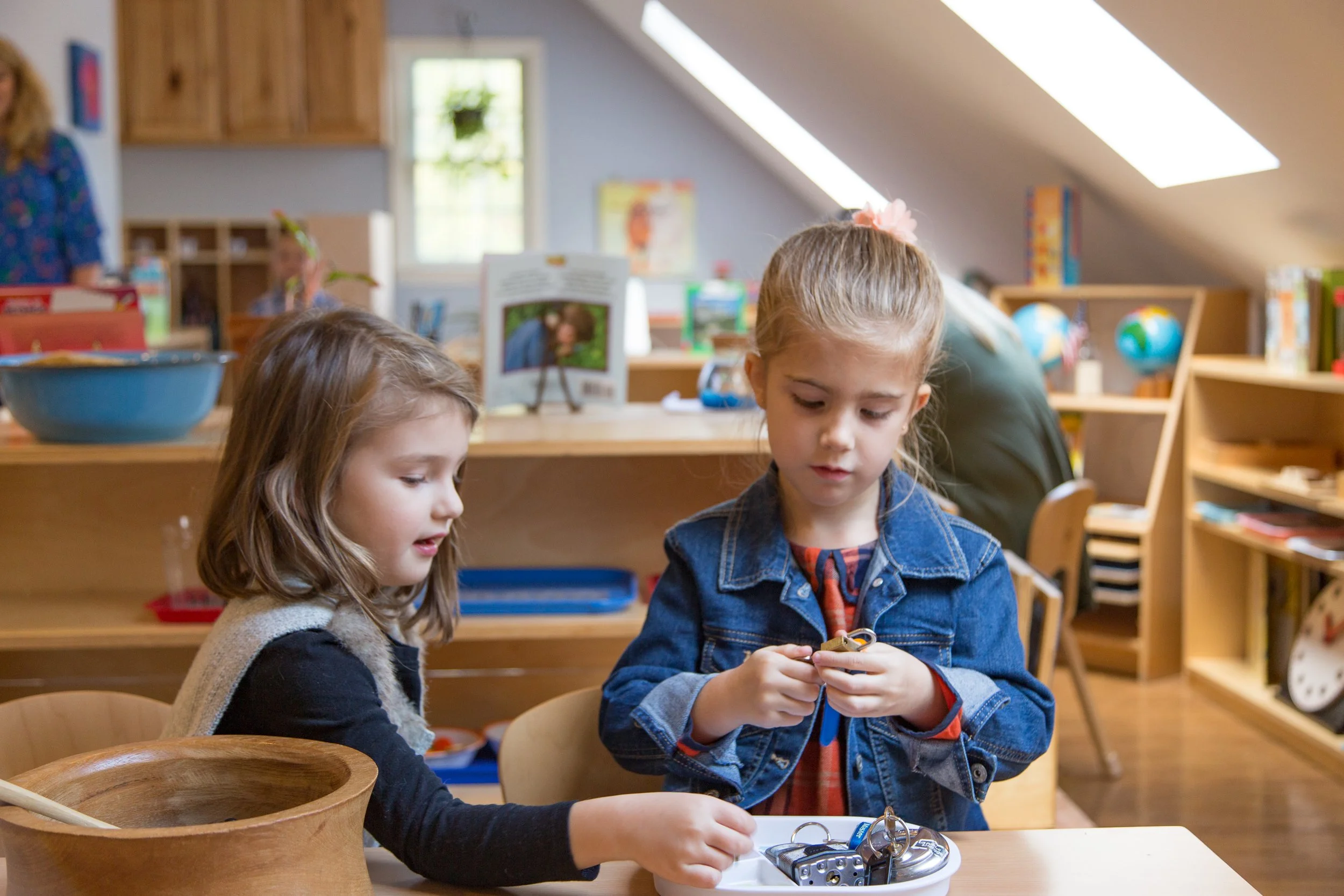 Two young girls sitting at a table in a classroom, examining keys and locks, with shelves and educational materials in the background.