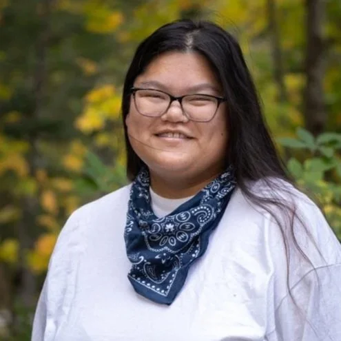 A woman with long dark hair and glasses smiling outdoors, wearing a white shirt and a blue bandana, with trees and yellow leaves in the background.