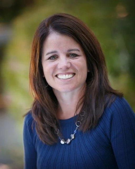 A woman with brown hair smiling outdoors, wearing a blue sweater and a silver necklace.
