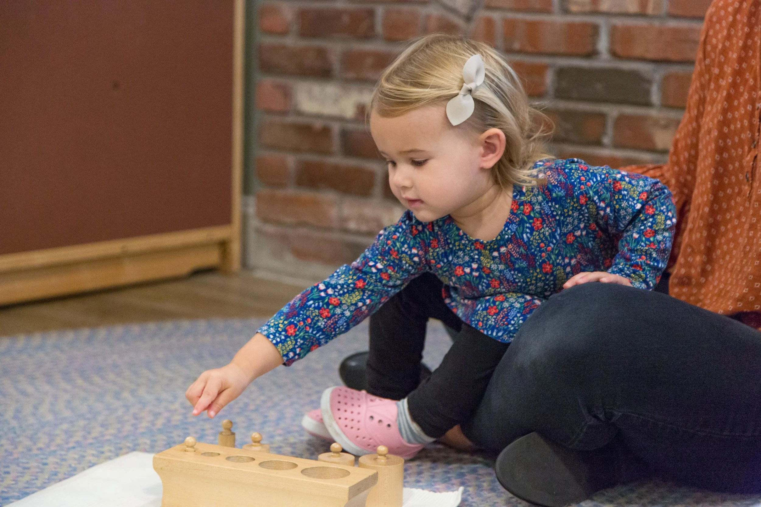 A young girl with a white hair bow playing a game with wooden pegs on the floor, sitting next to an adult, inside a room with brick wall and carpeted floor.