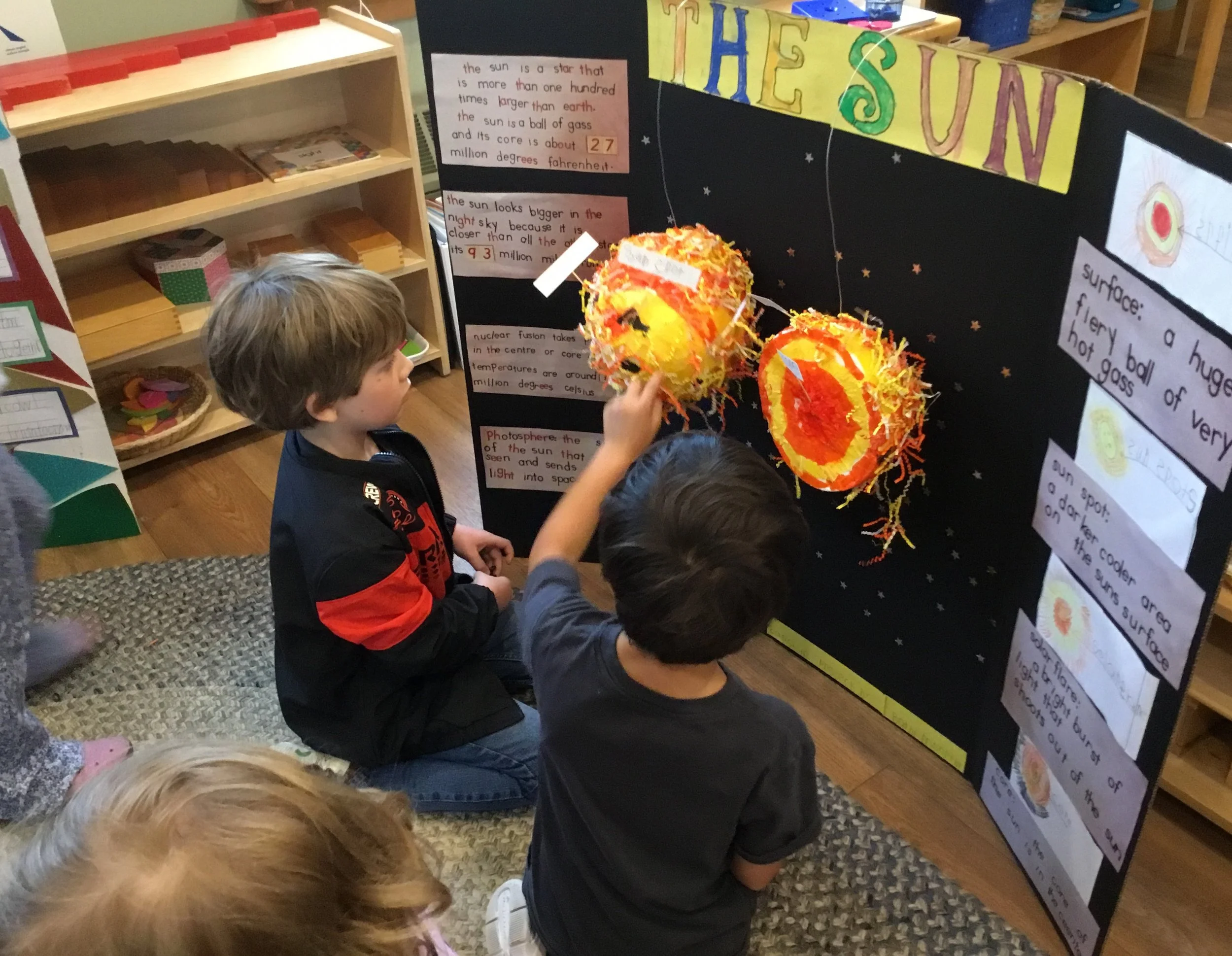 Children in a classroom interact with a science project display about the sun, which features a large paper mache sun with tissue paper flames and informational posters about the sun's characteristics.
