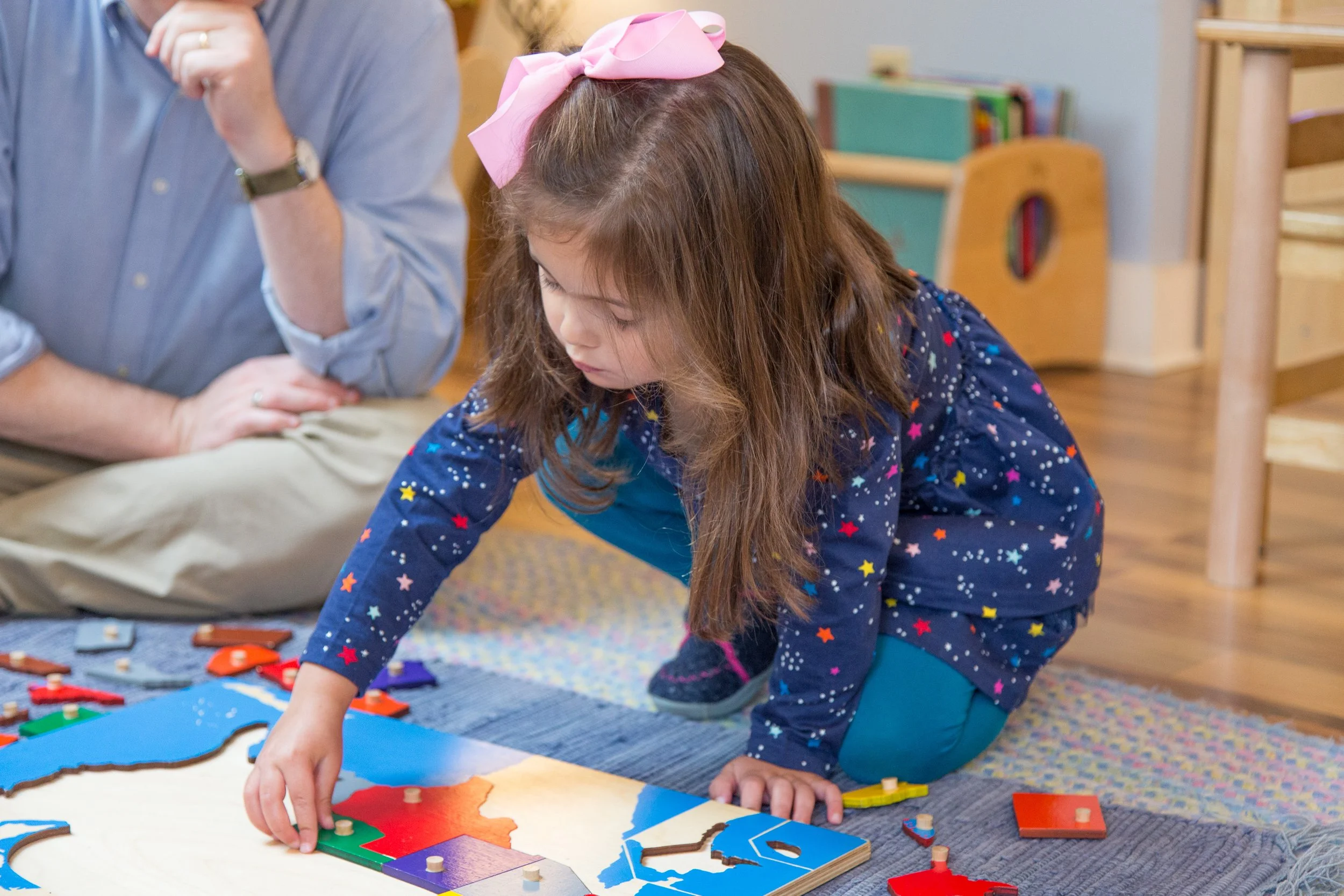 A young girl with long brown hair wearing a pink bow and a navy shirt with colorful stars, kneeling on the carpet, playing with a colorful wooden puzzle on a white table.