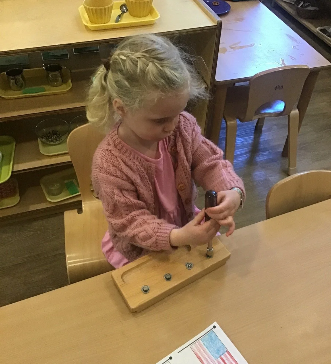 A young girl with blonde hair in a braid is playing with a small hammer on a wooden board with metal washers in a classroom.