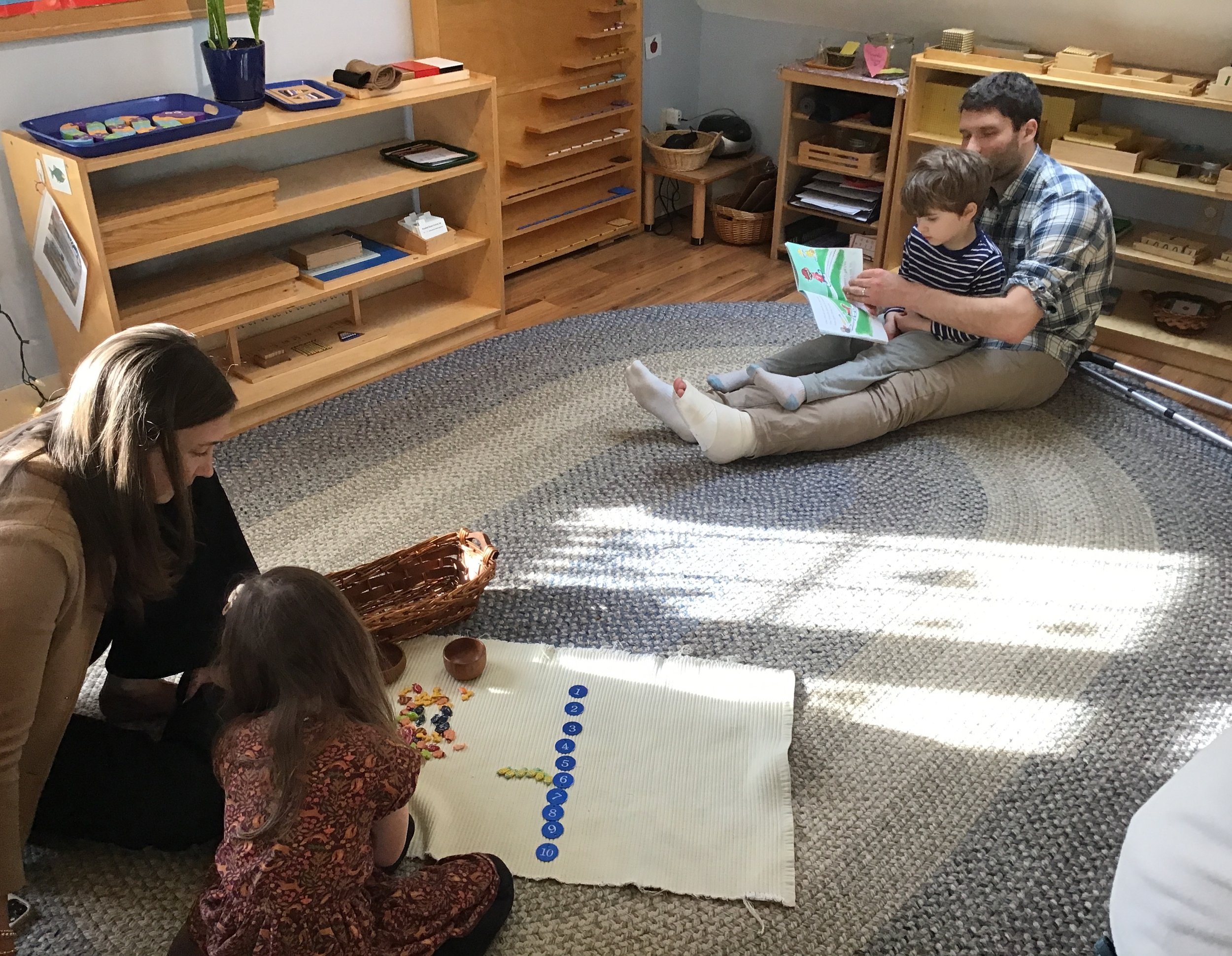 A young boy and his dad sit on the floor, reading a book together. Nearby, a mother and daughter are working on the floor together. The room has wooden shelves filled with educational materials and an elliptical, patterned rug.