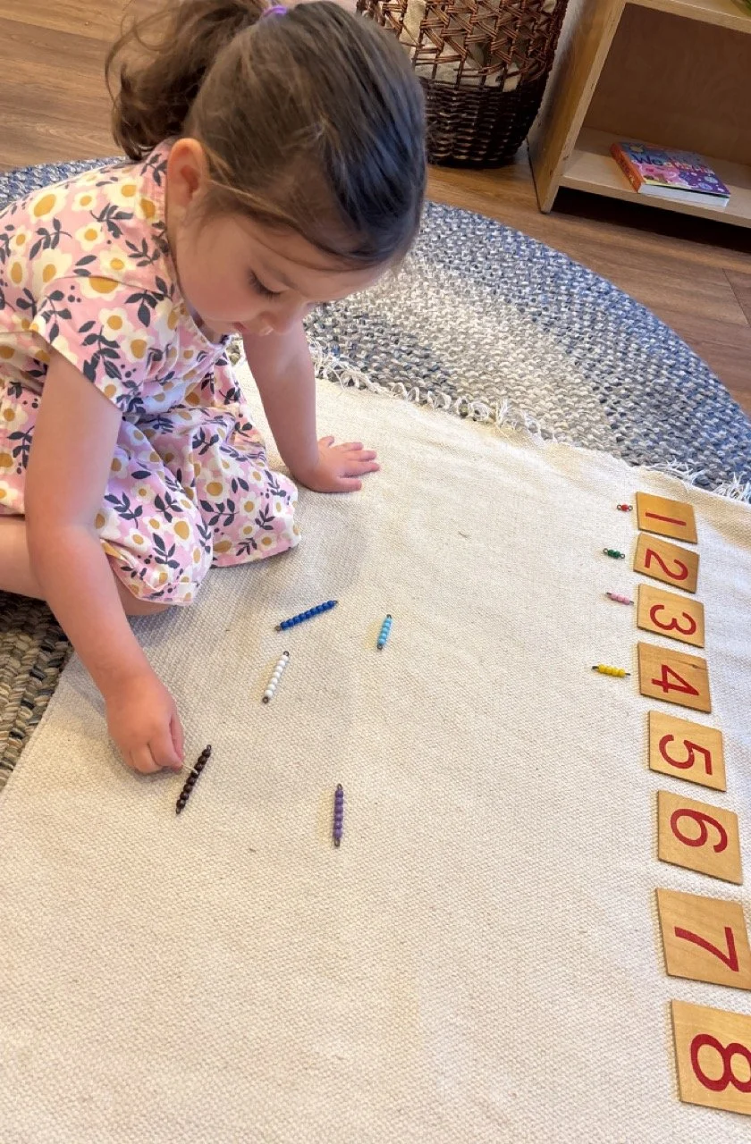 A young girl in a floral dress is sitting on the floor, engaged in counting or organizing small colored beads and number cards arranged in a sequence from 1 to 10 on a beige rug.
