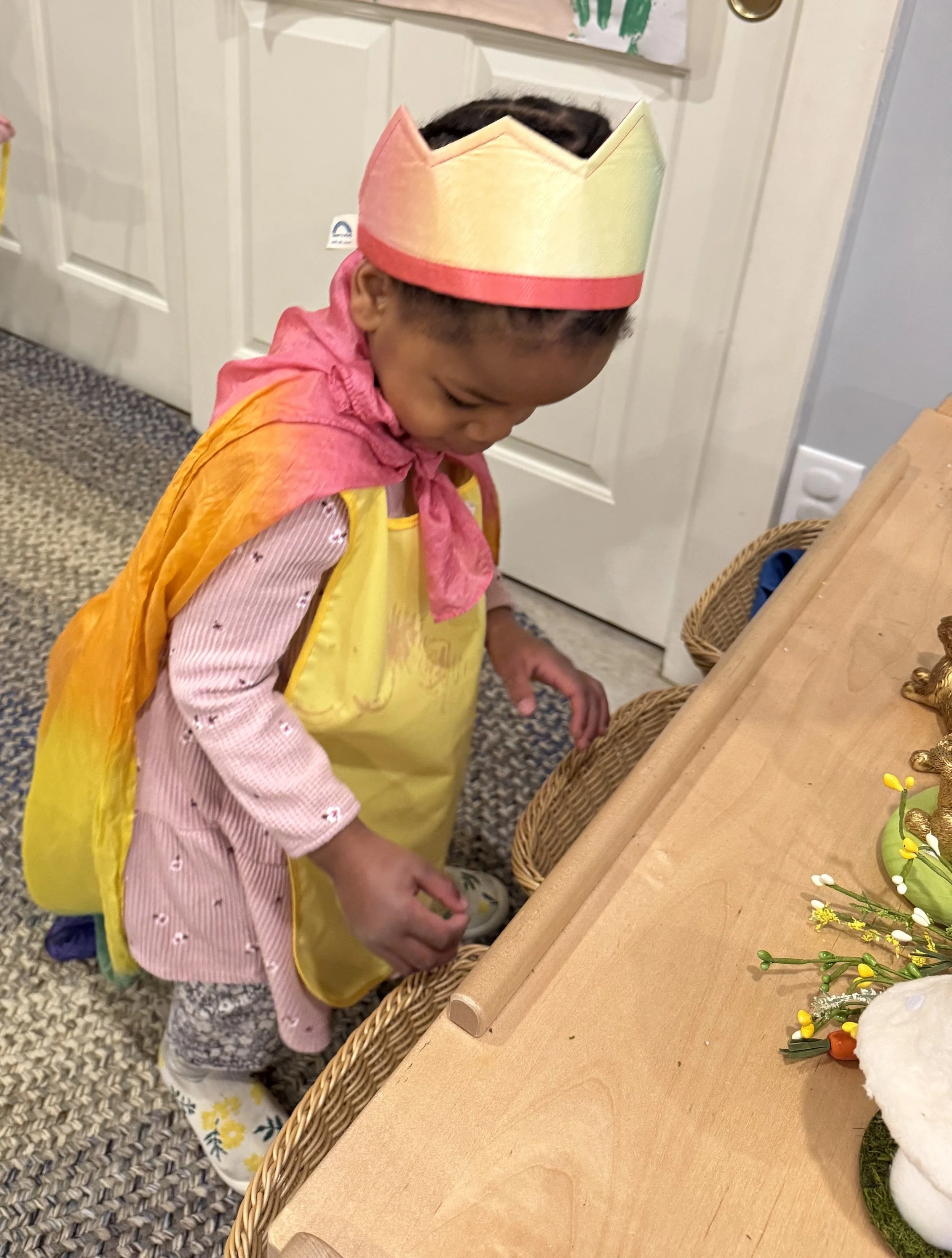 A young girl wearing a colorful paper crown, a yellow apron, and pink clothing with a pattern, standing by a table with baskets and decorations, looking down at something with her head slightly bowed.