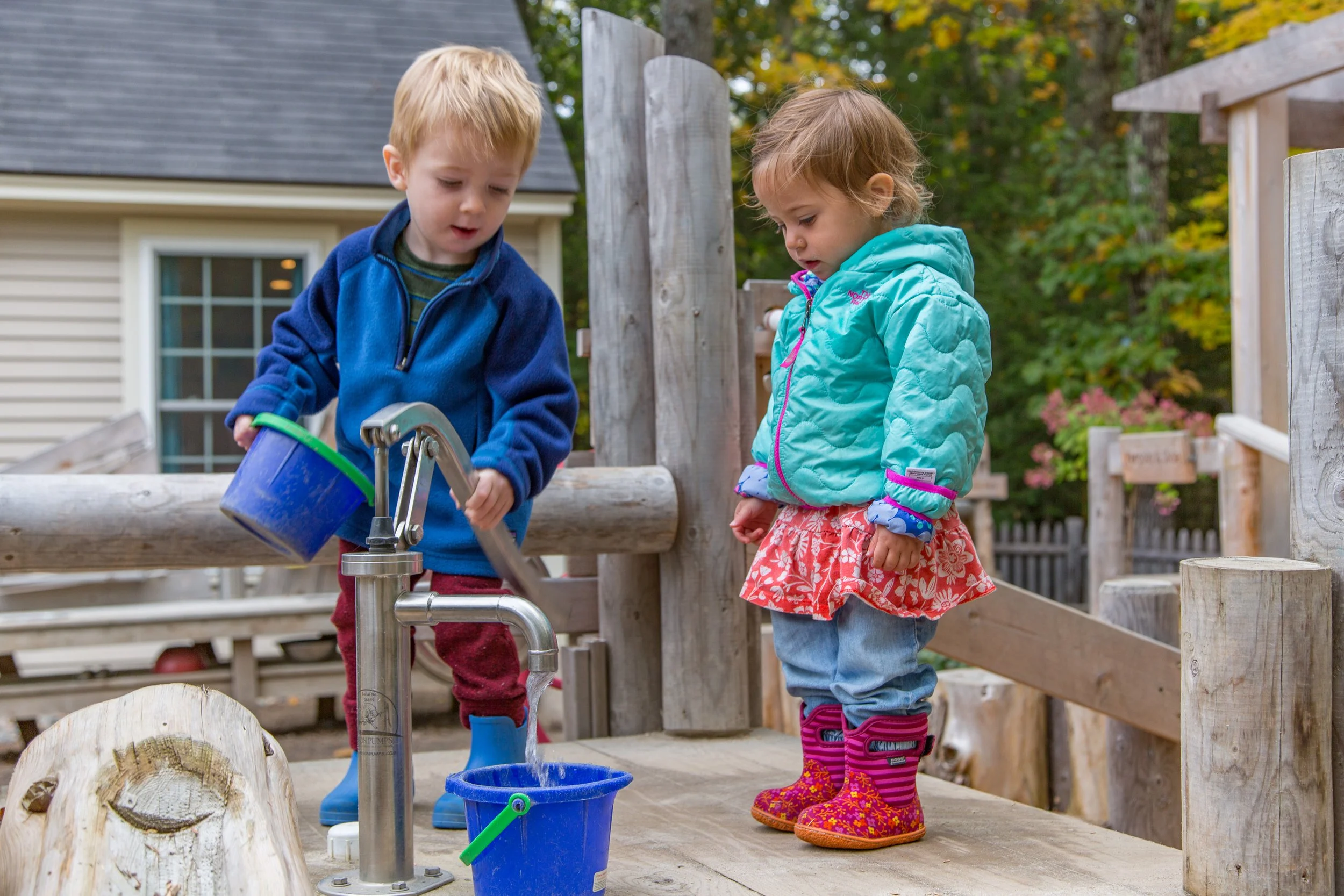 Two young children, a boy and a girl, playing at an outdoor water station. The boy is filling a blue container at a faucet, while the girl watches. They are dressed in colorful jackets and rain boots, with trees and a house in the background.