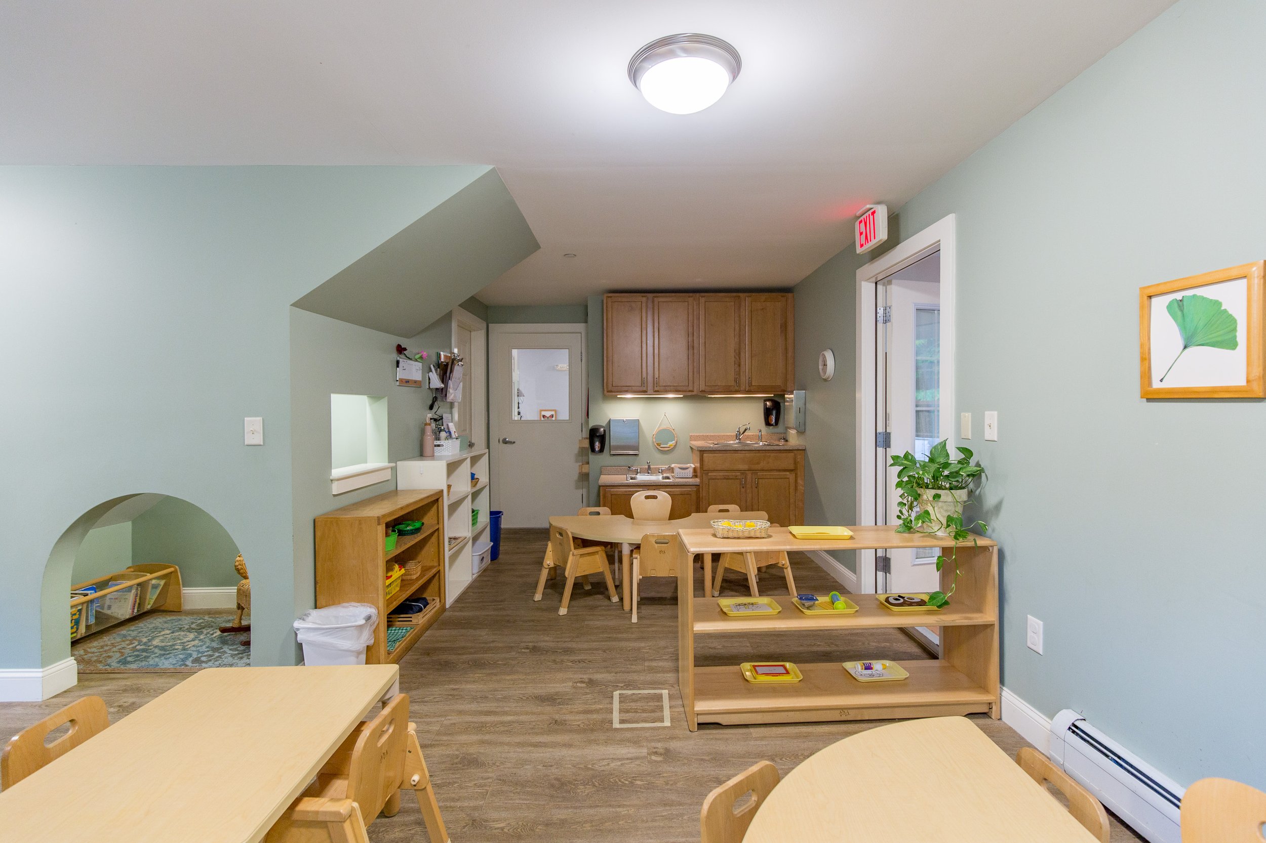 Interior of a children's classroom with small tables and chairs, wooden shelves, a kitchenette, and artwork on the walls.