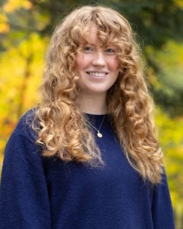 A young woman with curly red hair smiling outdoors, wearing a navy blue top and a gold necklace, with trees and yellow foliage in the background.