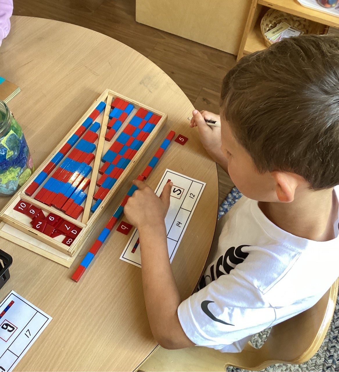 Child working on a math activity with red and blue counting blocks and number cards at a classroom table.