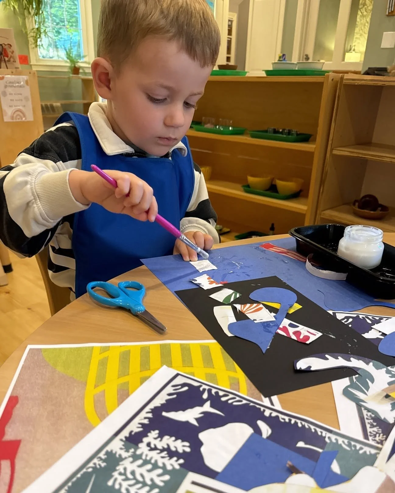 A young boy is sitting at a table, working on an arts and crafts project involving cutouts of a snowy landscape scene. He is using a pink glue stick and has a pair of blue scissors nearby. The background shows a room with wooden shelves and plates.