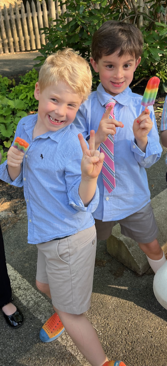 Two young boys smiling and posing outdoors, each holding a rainbow-colored popsicle and making peace signs with their hands. They are dressed in light blue shirts, with one boy wearing a striped tie and the other boy wearing beige shorts.