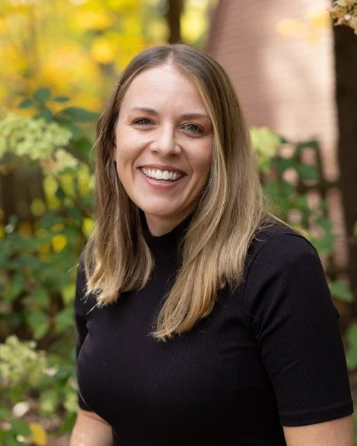 Smiling woman with long blonde hair wearing a black shirt outdoors with autumn foliage in the background.