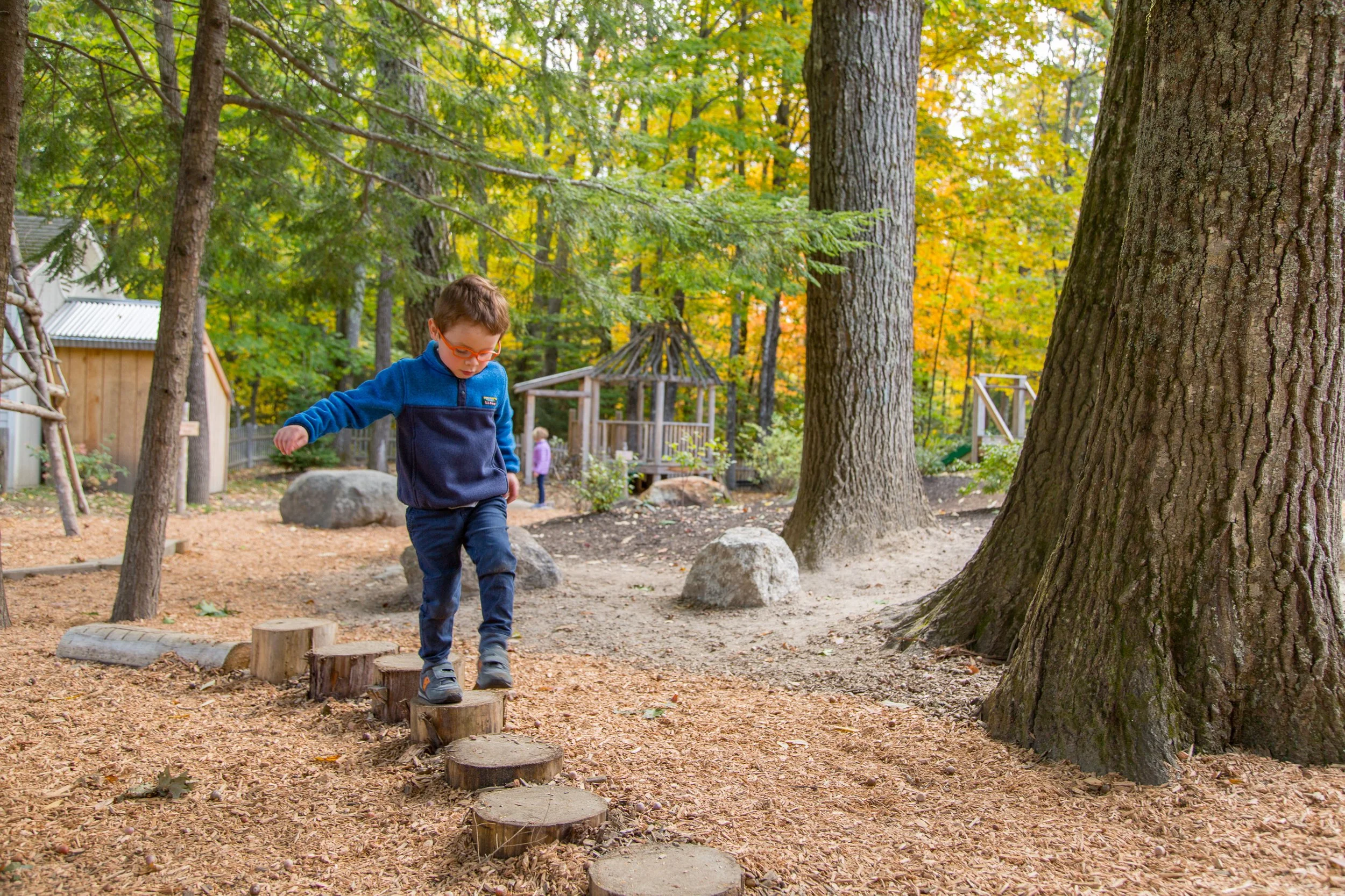 Young boy in a blue jacket and glasses balancing on wooden stepping stones in a wooded outdoor play area with trees, rocks, and other children in the background.
