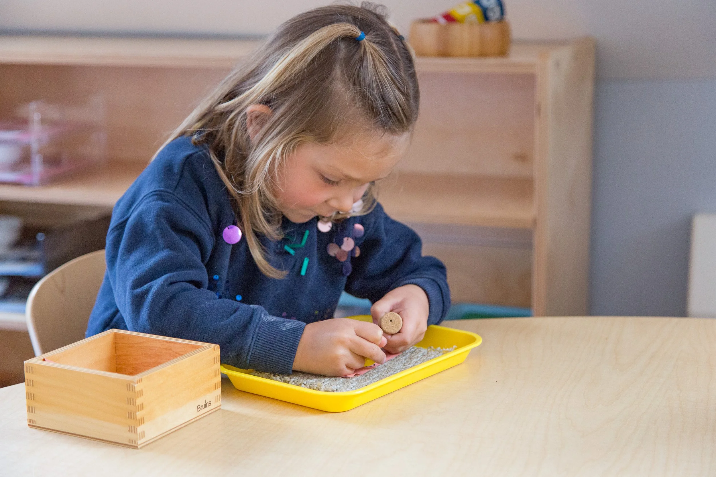 A young girl with blonde hair tied with a blue hairband, wearing a navy blue shirt, sitting at a wooden table, playing a game involving small cork pieces and a yellow tray. There are wooden boxes on the table, one labeled 'Bruins'. In the background, there is a wooden shelf with colorful objects.