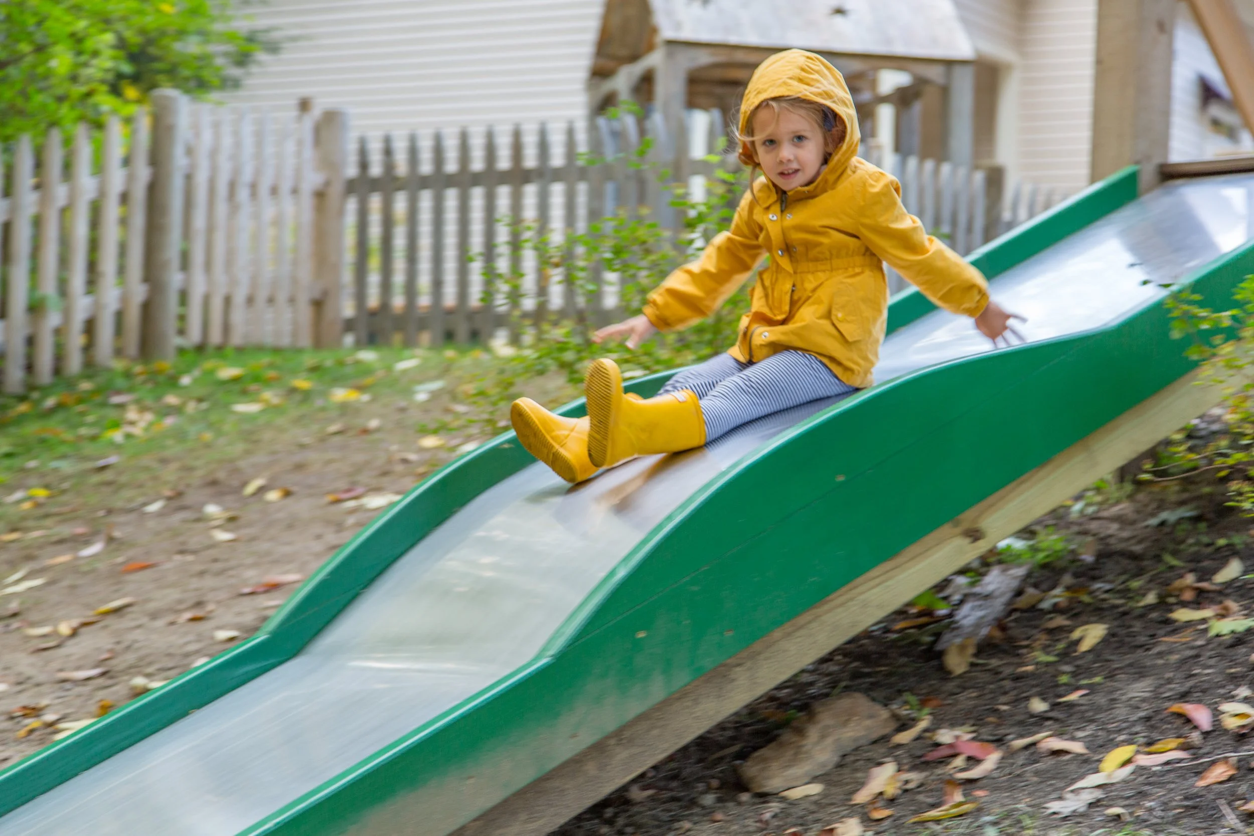 A young girl in a yellow raincoat and yellow rain boots sitting on a slide outdoors in a backyard with a fence and a house in the background.