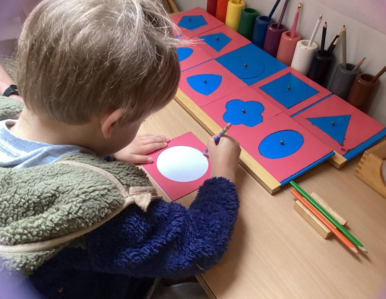 A young boy with light brown hair wearing a gray hoodie and a navy fuzzy jacket, sitting at a wooden table, coloring a white circle on a red card. In front of him are boards with red backgrounds and blue shapes, resembling geometric figures, each with a small metal knob in the center. Behind the boards are containers holding colored pencils and markers.