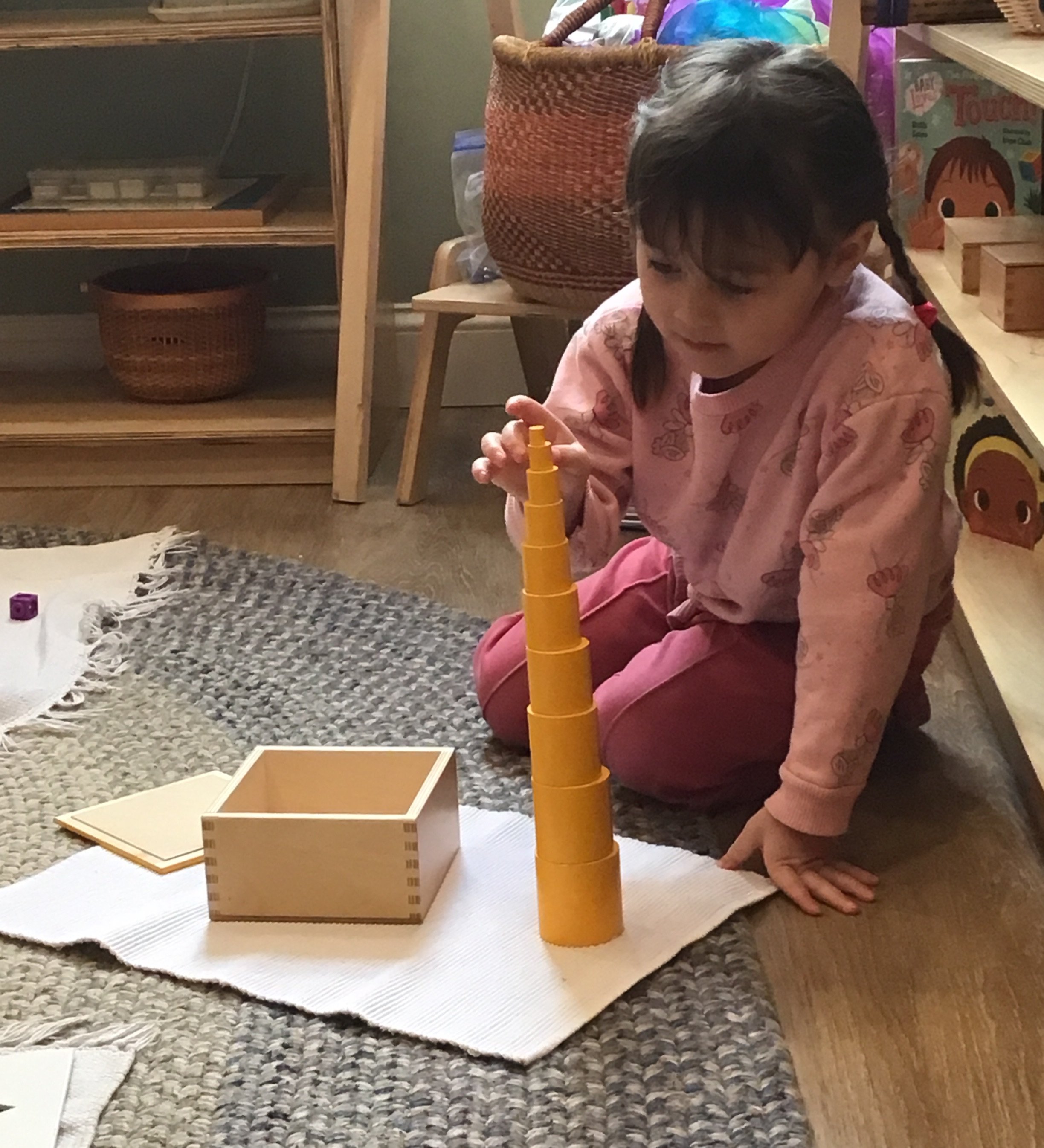A young girl with braided hair sitting on the floor, playing a stacking game with yellow pieces. There are wooden boxes and shelves with toys and books in the background, in a cozy indoor setting.