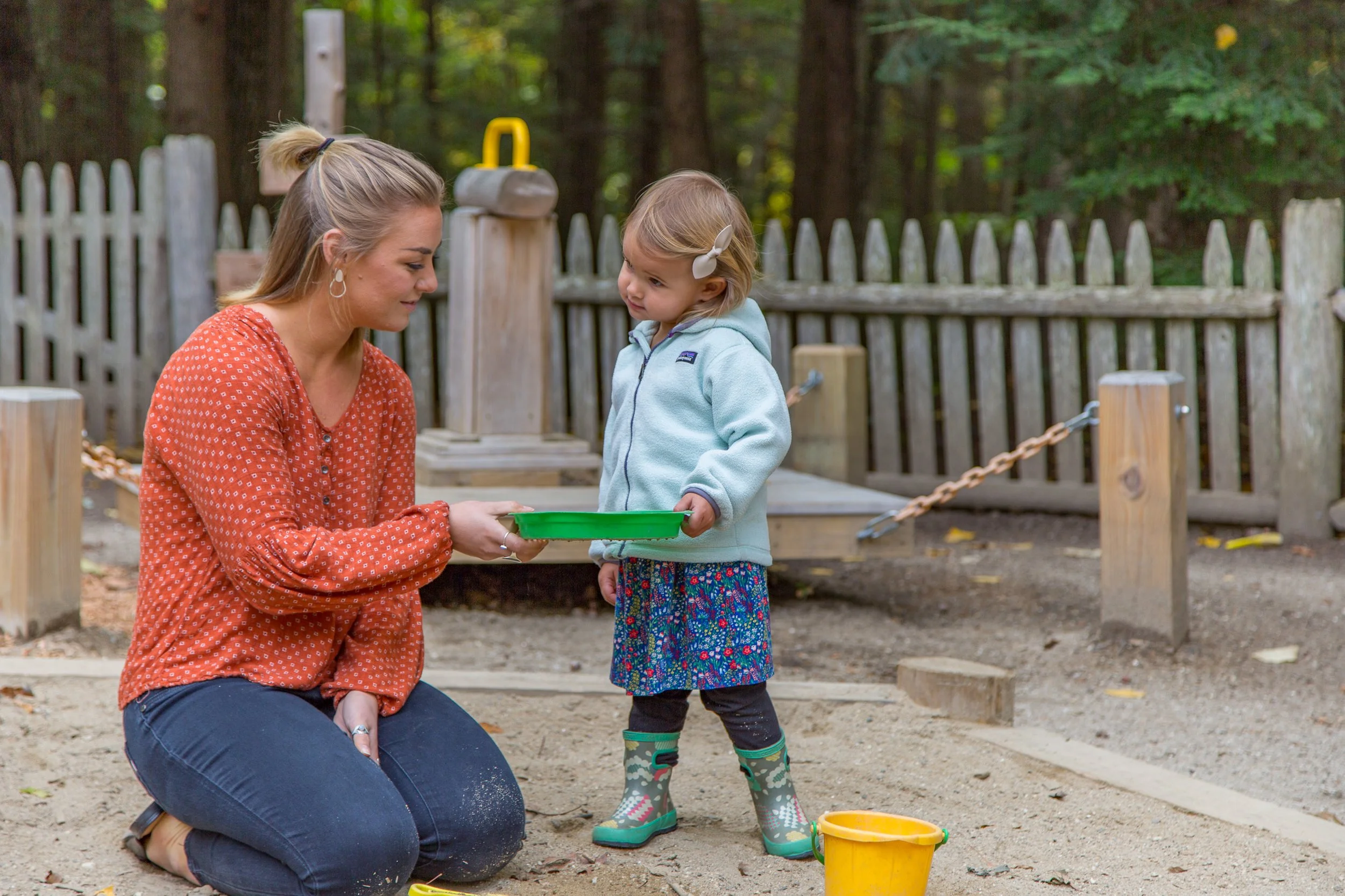 A woman kneeling in the sand with a young girl standing next to her at a playground. The woman is holding a green tray and the girl is about to take it. The girl is wearing a light blue hooded jacket, a colorful skirt, black leggings, and green rain boots. There is a yellow bucket on the ground nearby, and the background includes a wooden fence, trees, and playground equipment.