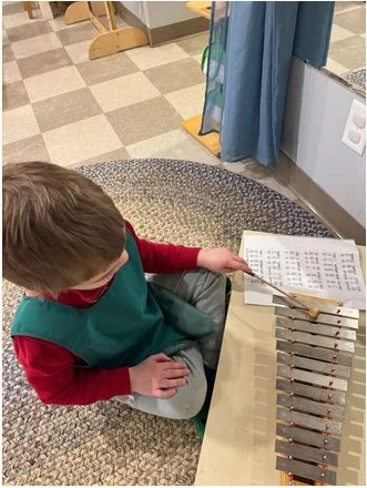 A young boy sitting on the floor with a music xylophone in front of him, holding a sheet of paper and a pencil in a classroom or music room.
