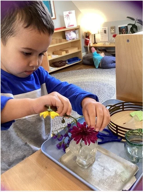A young boy arranging colorful flowers in a small glass vase on a tray at a table.
