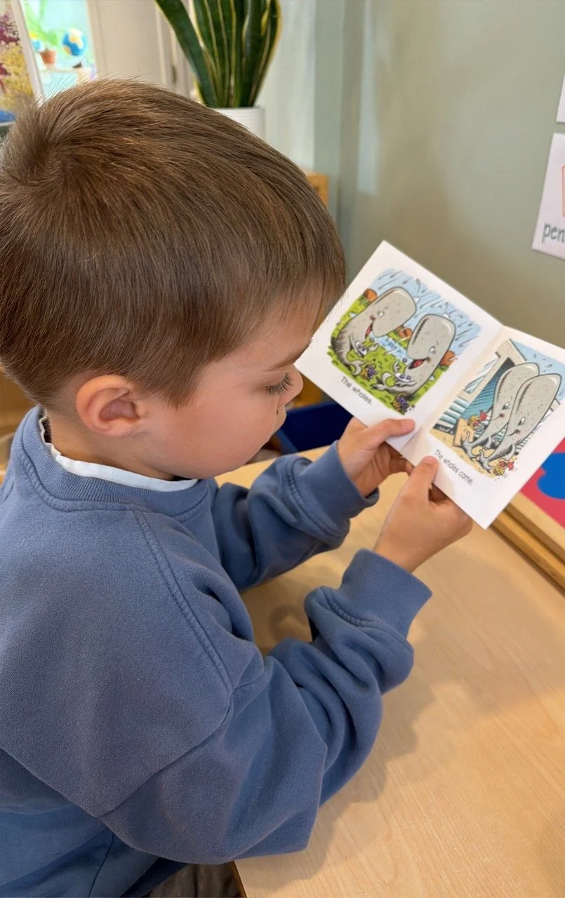 A young boy with light brown hair reading a small open book with cartoon characters named inside, sitting at a wooden table in a classroom setting.