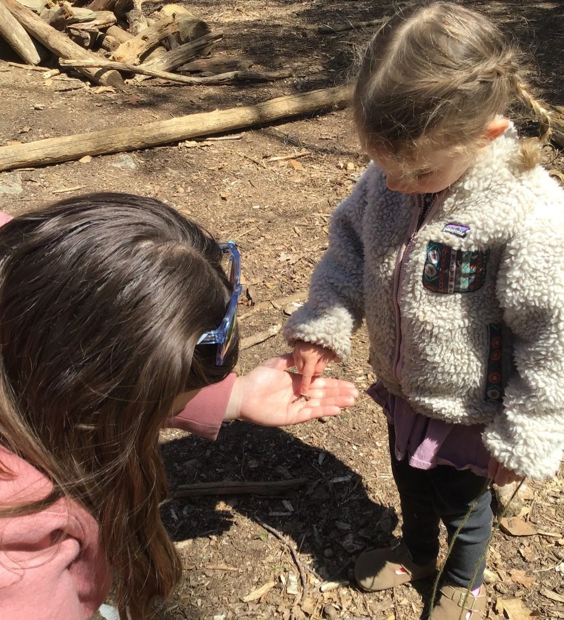 Two people outdoors, one crouching and holding a small worm in her palm, the other looking down at her hand, wearing a warm fleece jacket.