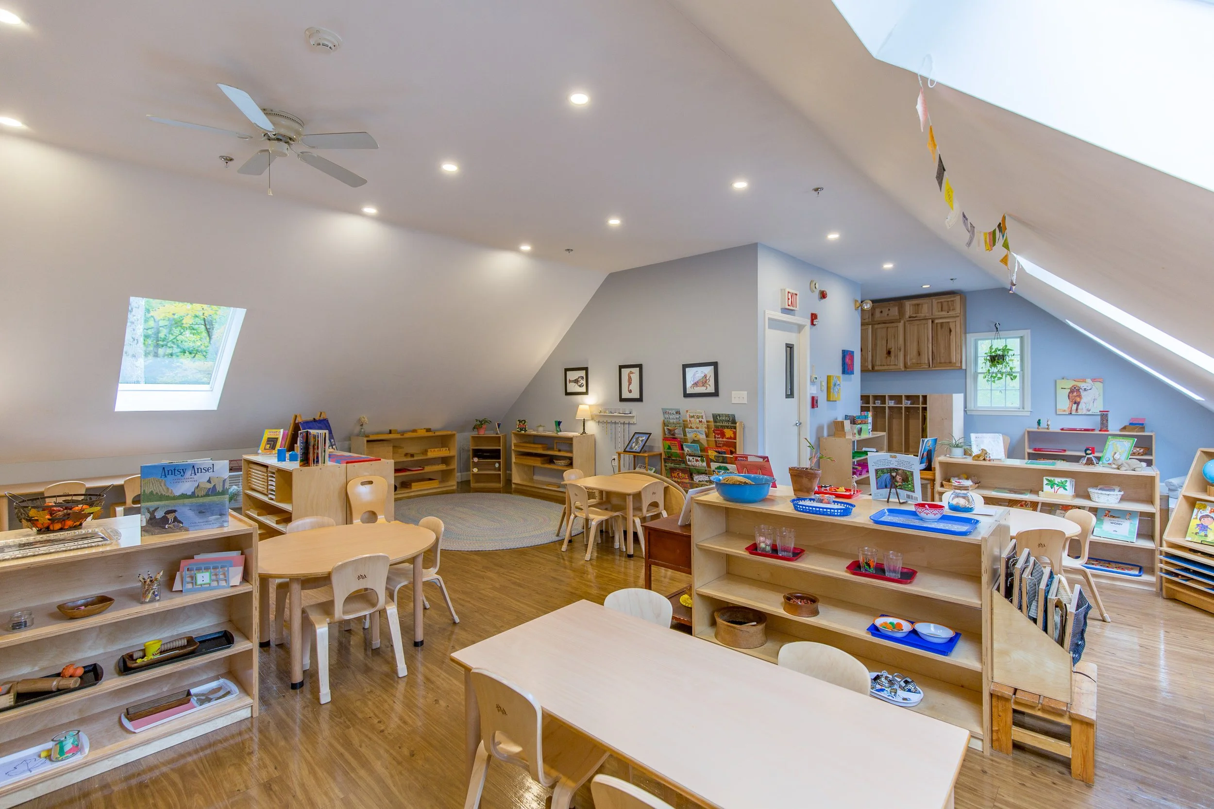 A spacious, well-lit classroom with sloped ceiling, skylights, and wooden floors, furnished with small tables, chairs, and shelves filled with books and educational materials, decor includes framed pictures on the wall and a colorful banner hanging from the ceiling.