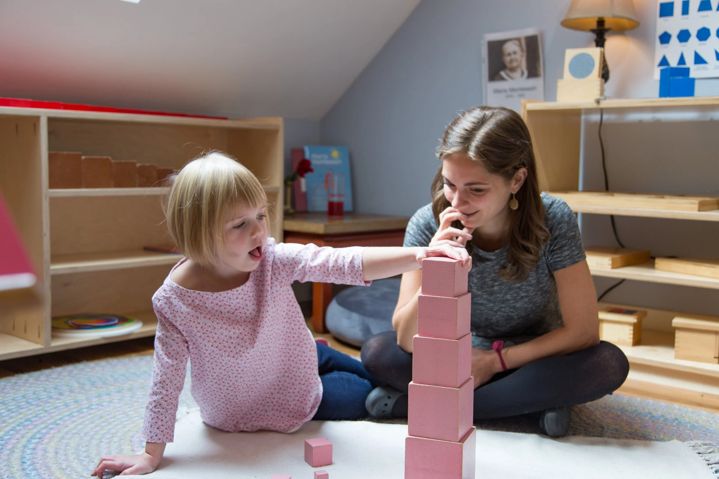 A young girl is working with the Pink Tower in a Montessori classroom. The girl is reaching out to touch the cubes, and the teacher is watching her, smiling.