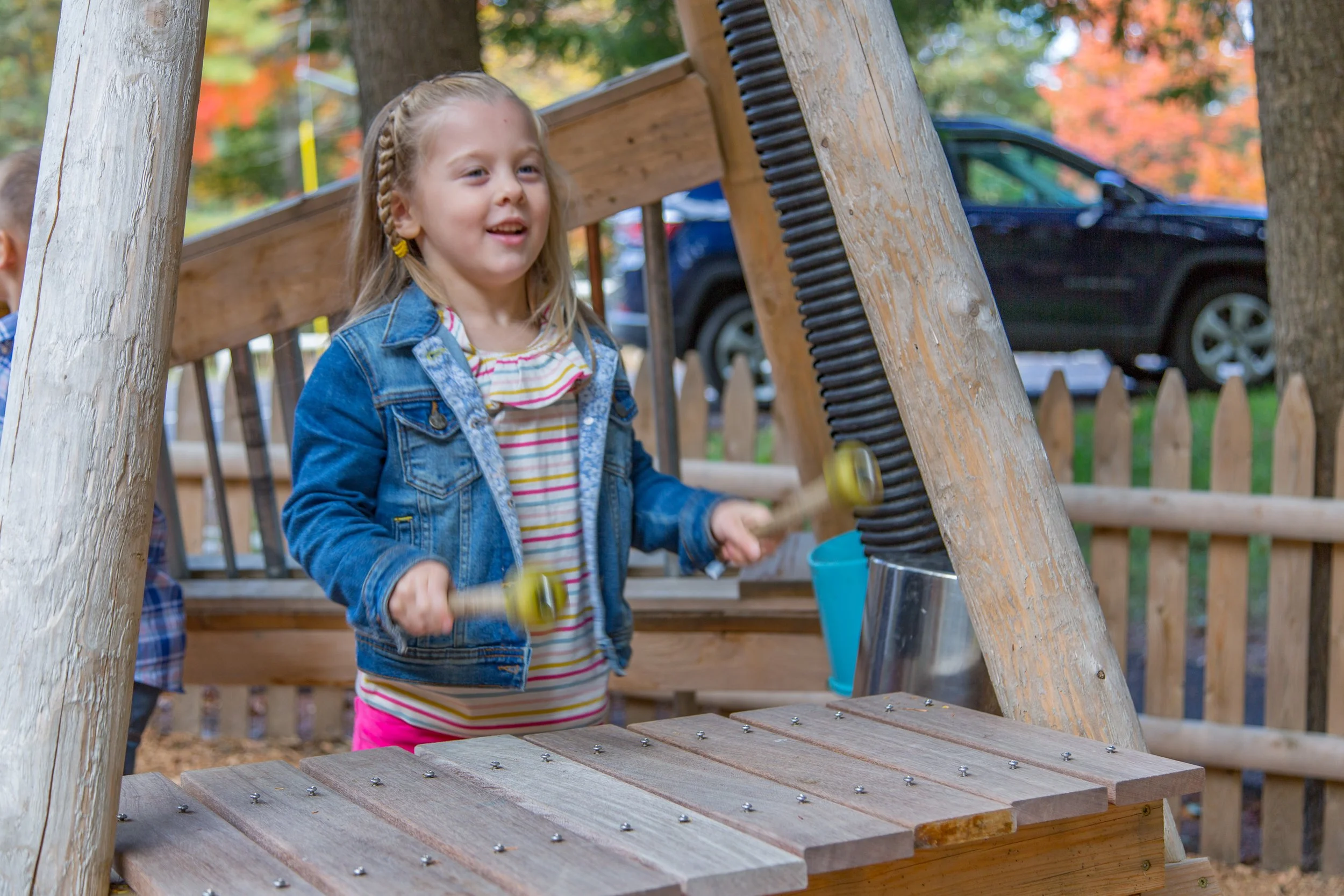 A young girl with braided hair wearing a denim jacket and pink pants playing on a wooden play structure in a park with autumn-colored trees in the background.