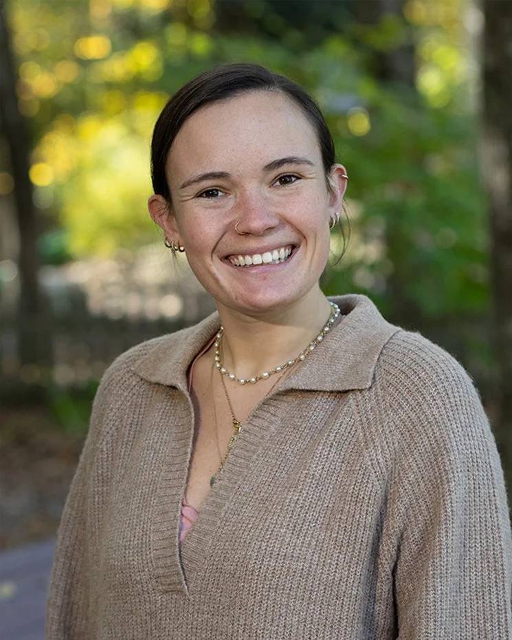 A young woman smiling outdoors on a sunny day, wearing a beige sweater, multiple necklaces, and earrings, with trees and sunlight in the background.