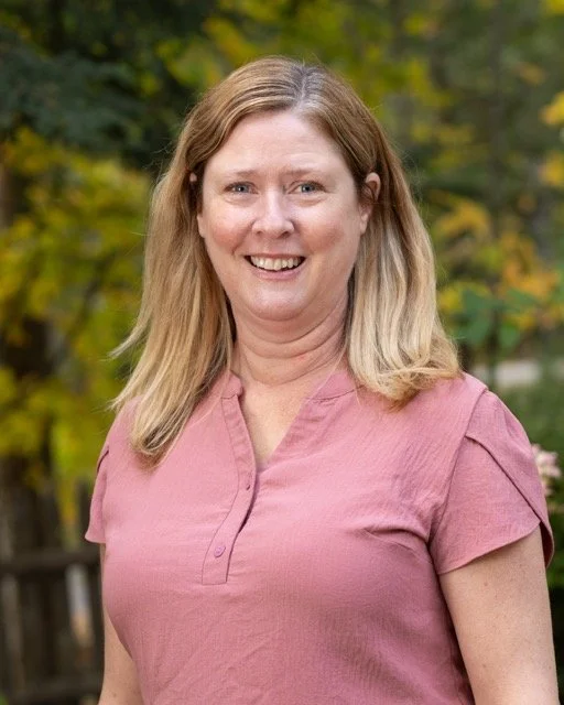 A smiling woman with blonde hair wearing a pink shirt outdoors with green and yellow trees in the background.