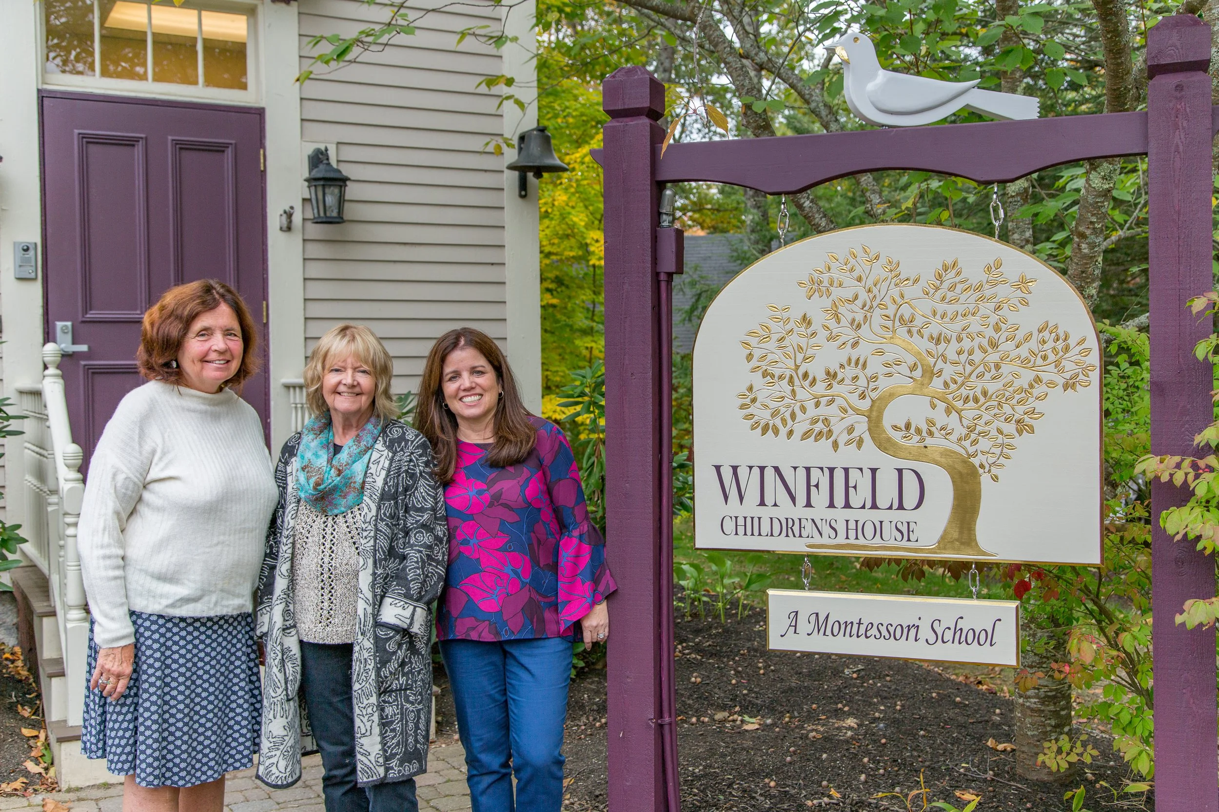 Three women standing outside in front of a house, smiling, next to a sign that reads 'Winfield Children's House - A Montessori School' with a tree logo and a white bird ornament on the purple wooden frame.