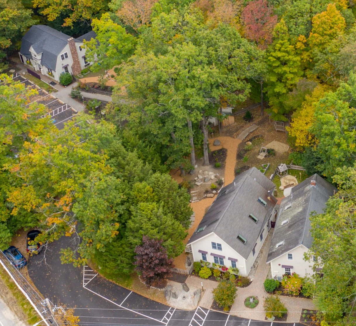 Aerial view of residential houses surrounded by trees with fall foliage, parking lot, and pathways in a suburban neighborhood.