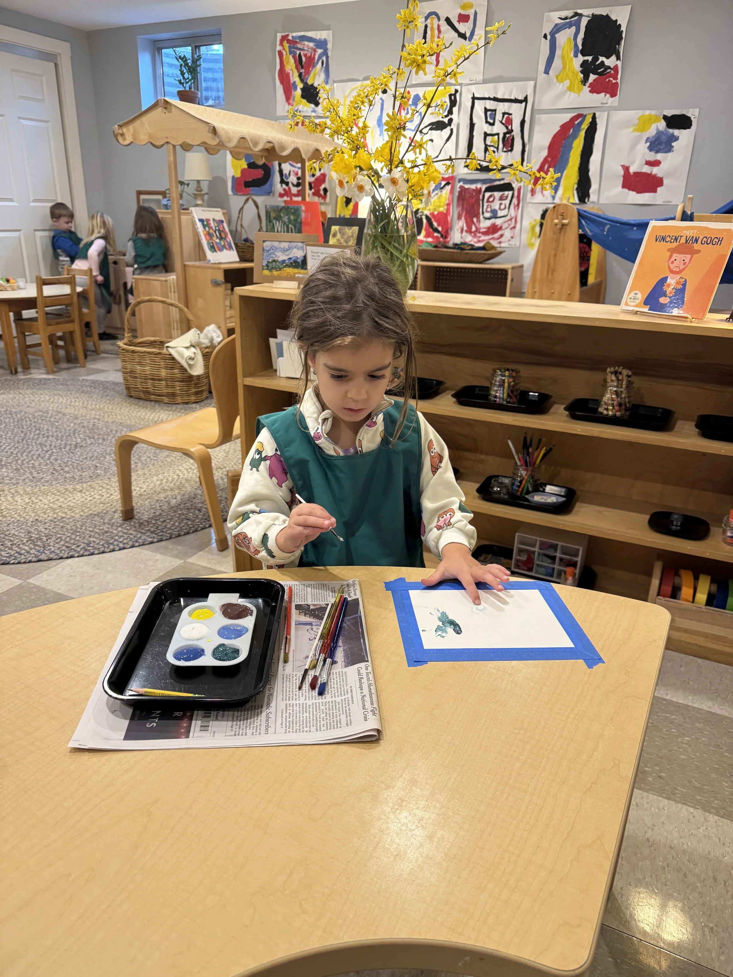 A young girl working on a watercolor painting at a table in a classroom or art room, with art supplies, a newspaper, and her paper taped down. In the background, a vase with yellow flowers and watercolor paintings on the wall are visible.