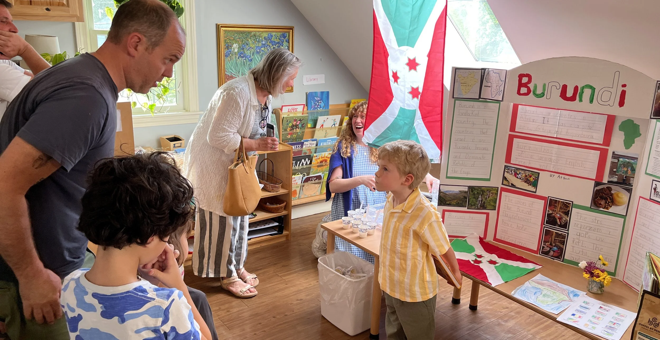 Children and adults at a school fair or presentation, with a large Burundi flag and a poster about Burundi on display.