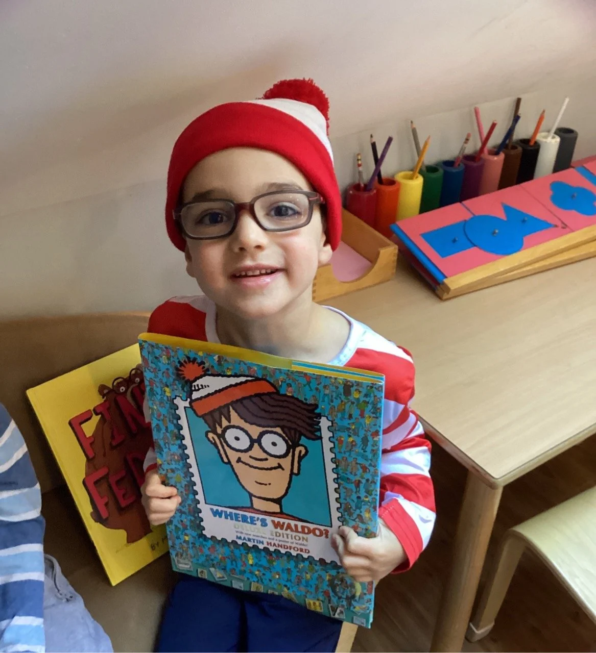 A young boy wearing glasses and a red and white winter hat holding a "Where's Waldo" book, sitting next to a wooden table with colorful art supplies and a child's drawing.
