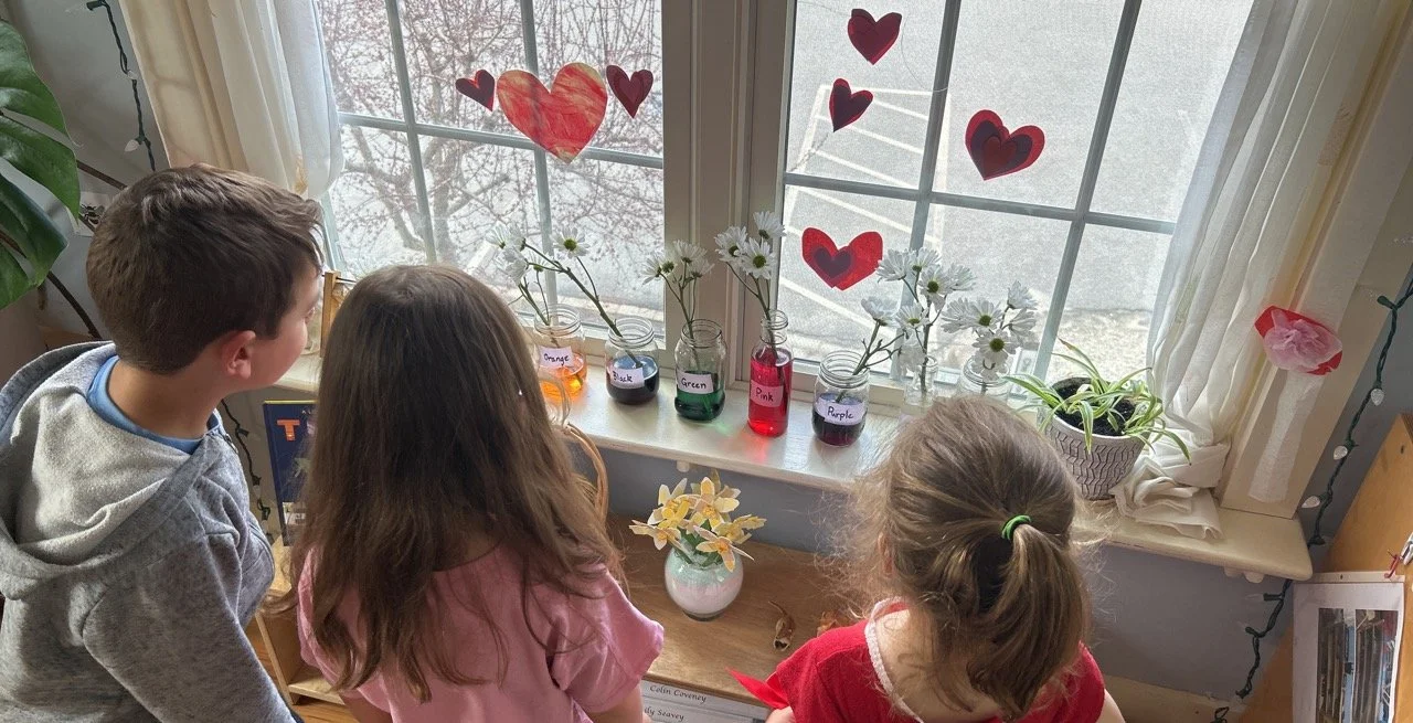 Three children looking out a window decorated with paper hearts, with vases of flowers and jars of colored water on the windowsill.
