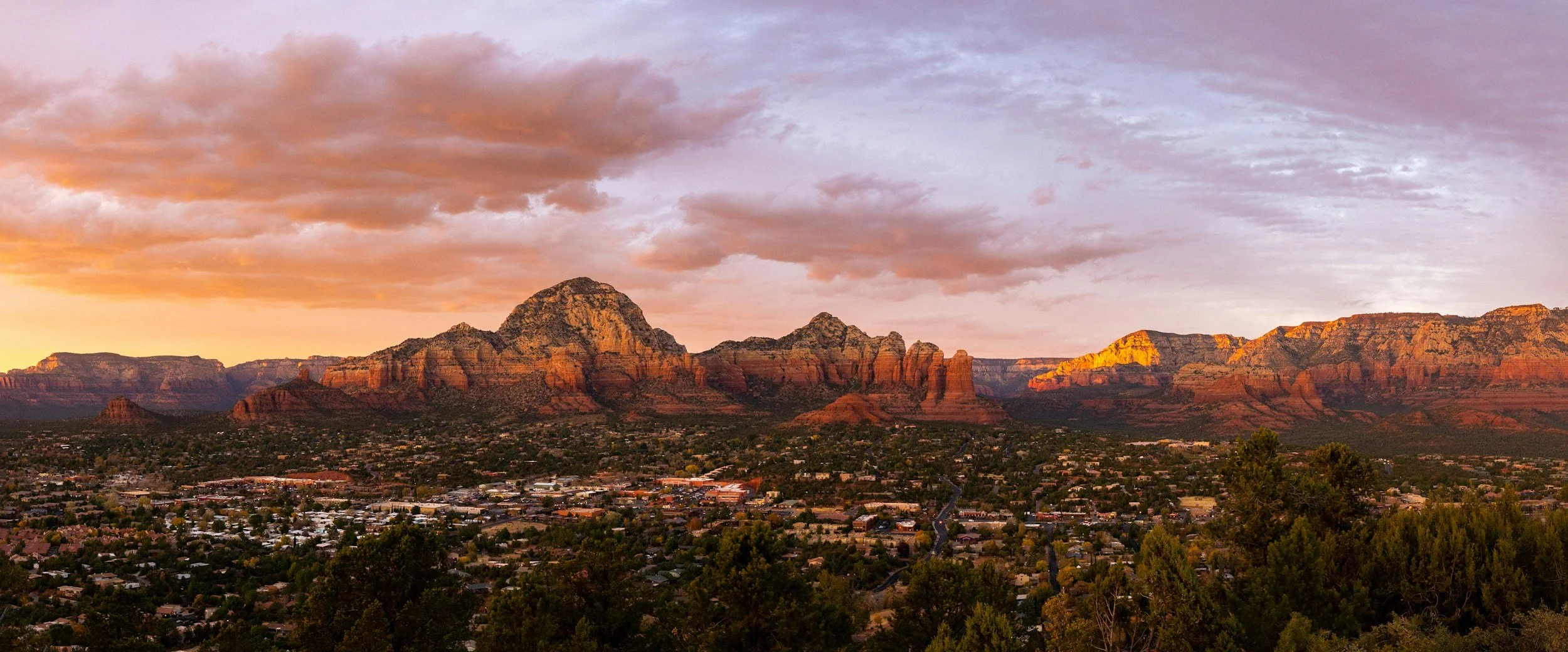 Sunset over red rock mountains with a city in the foreground and a cloudy sky.