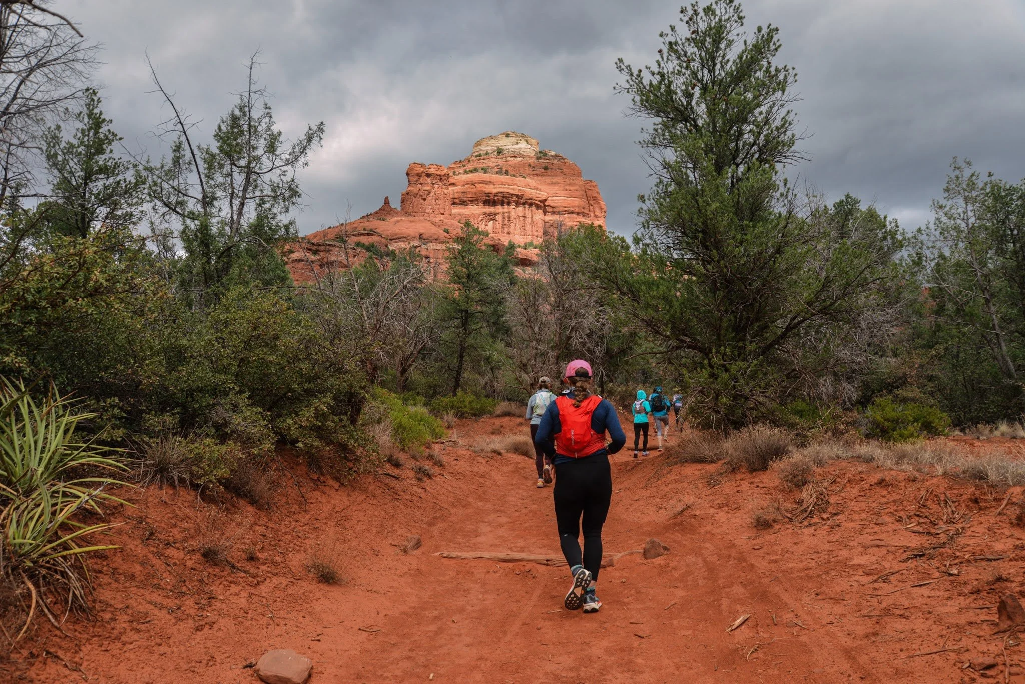 Group of runners walking on a trail surrounded by trees and red rocks, with a large rock formation and cloudy sky in the background.
