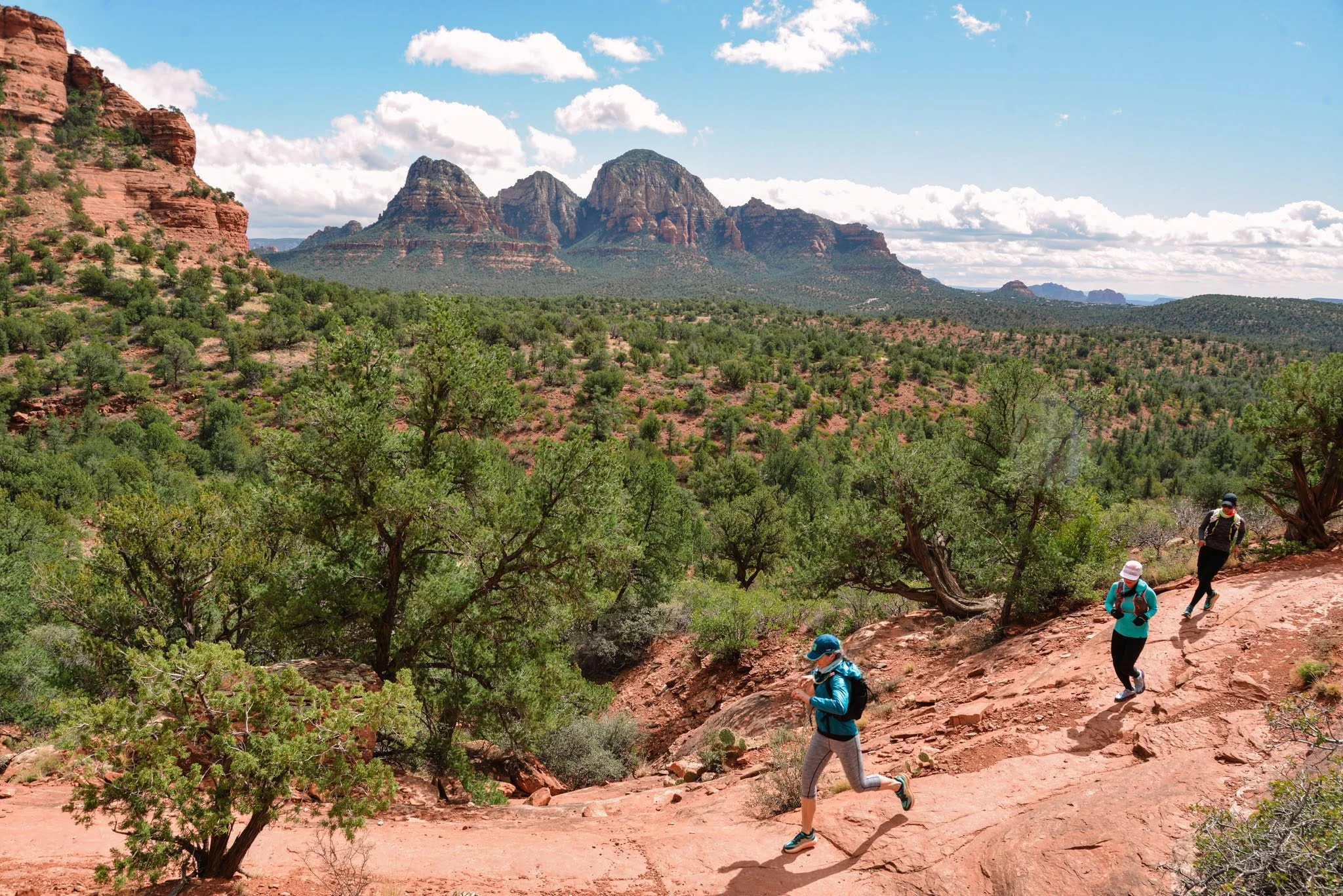 Four runners trekking on a reddish trail in a desert landscape with green trees and distant red rock formations and mountains under a partly cloudy sky.