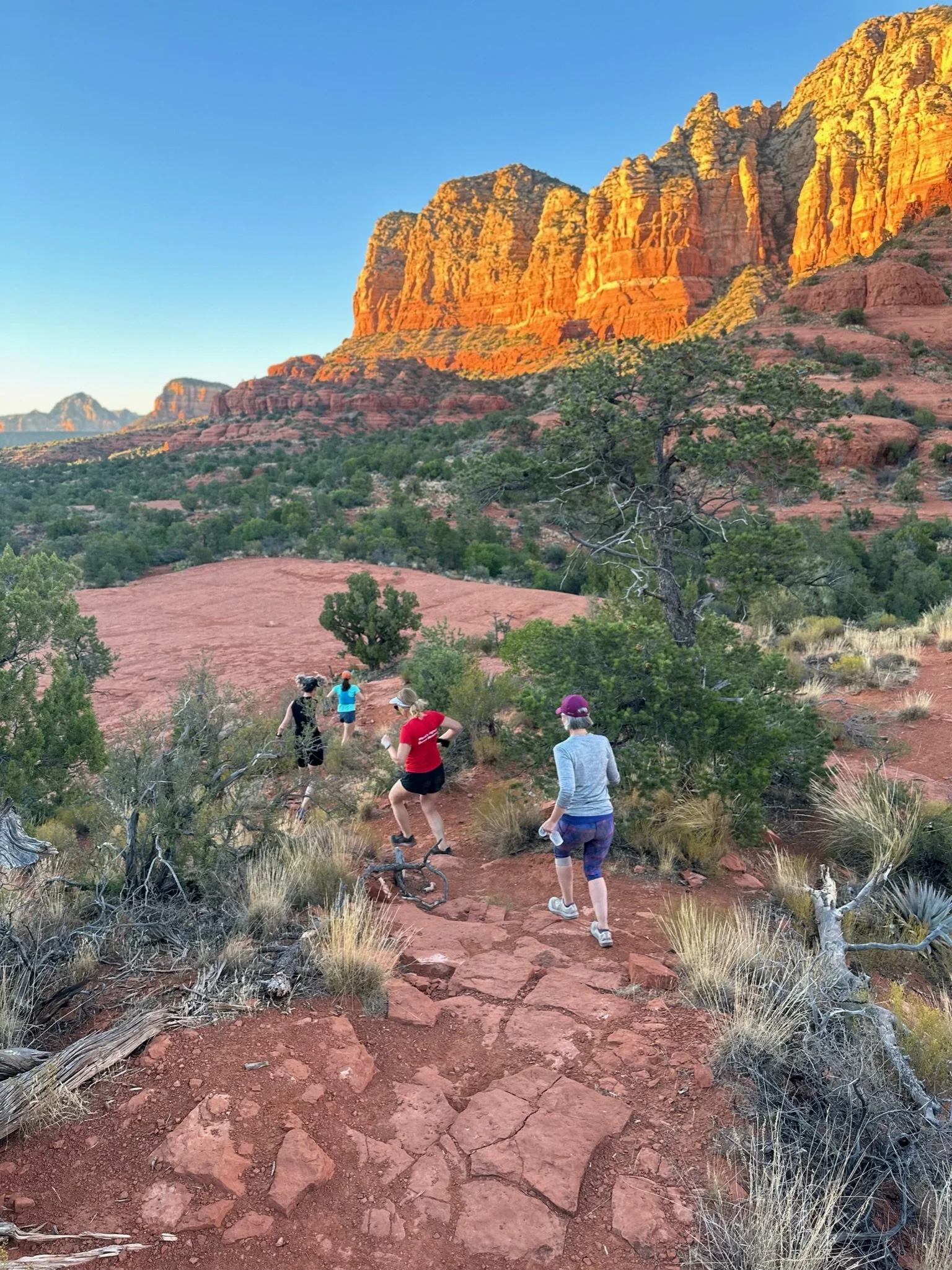 Group of people runningon a dirt trail through desert landscape with red rocks and cliffs in the background at sunset.