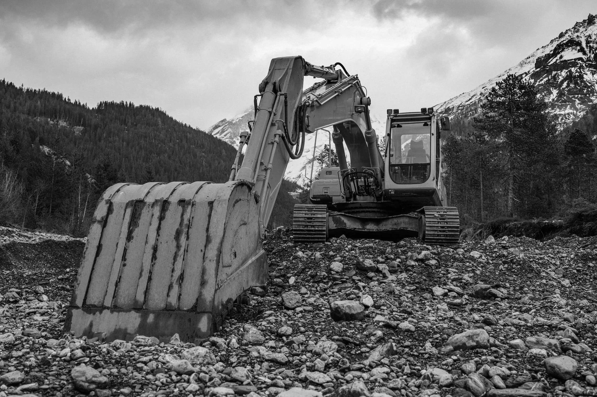 Black and white photo of an excavator on rocky terrain with mountains in the background.